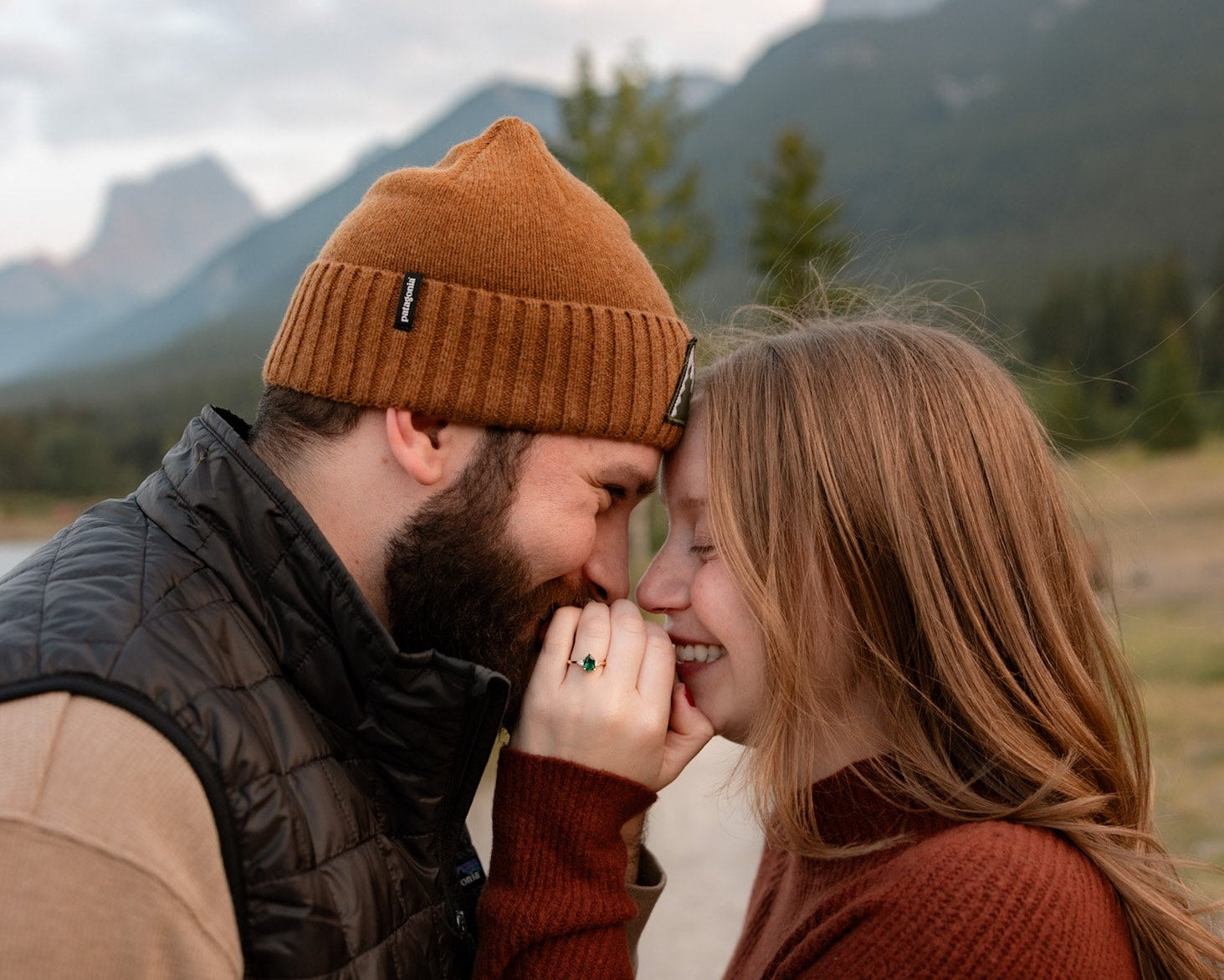 Couple in warm clothing embracing in Canmore, Alberta with mountains in the background and showing an emerald custom engagement ring on the woman's ring finger.