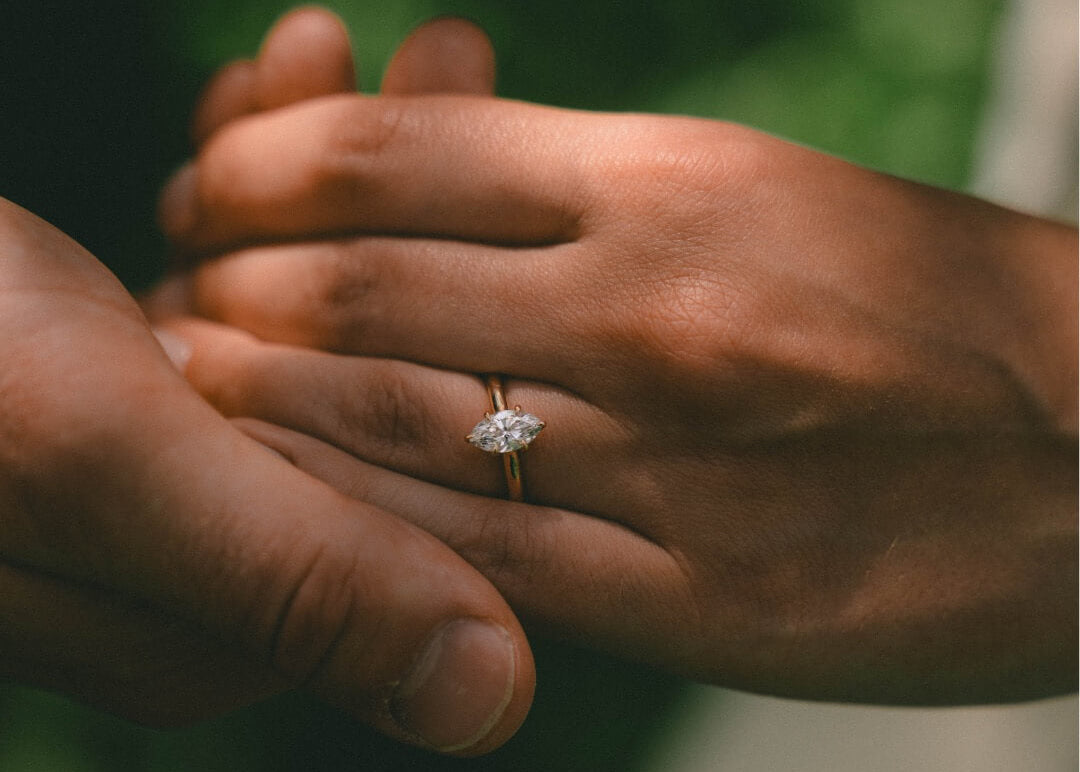 Close-up of two hands holding each other with a diamond ring on one finger, against a blurred green background.